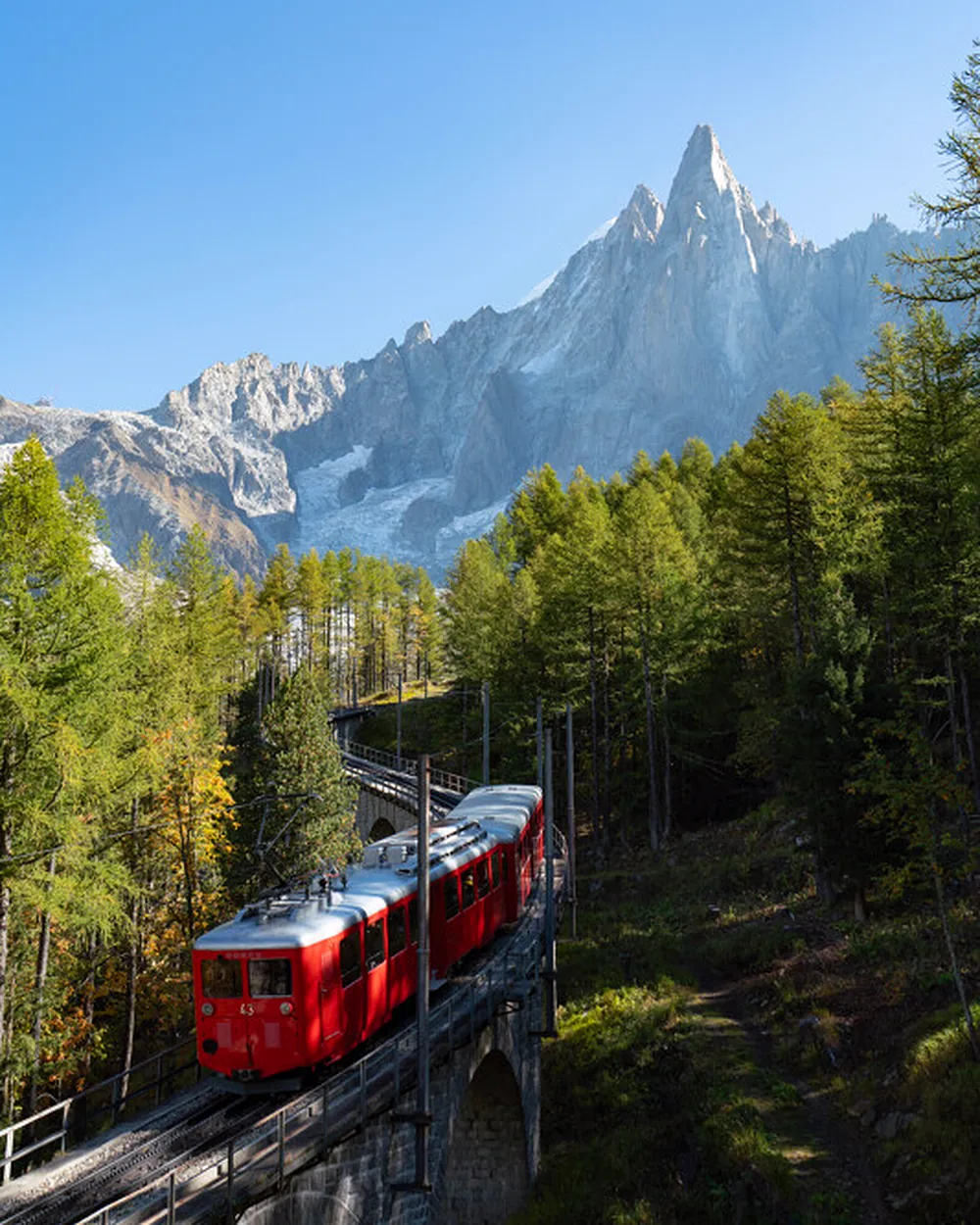 The red Montenvers railway train crossing a stone arch bridge through autumn larches, with the granite peaks of the Mont Blanc massif beyond